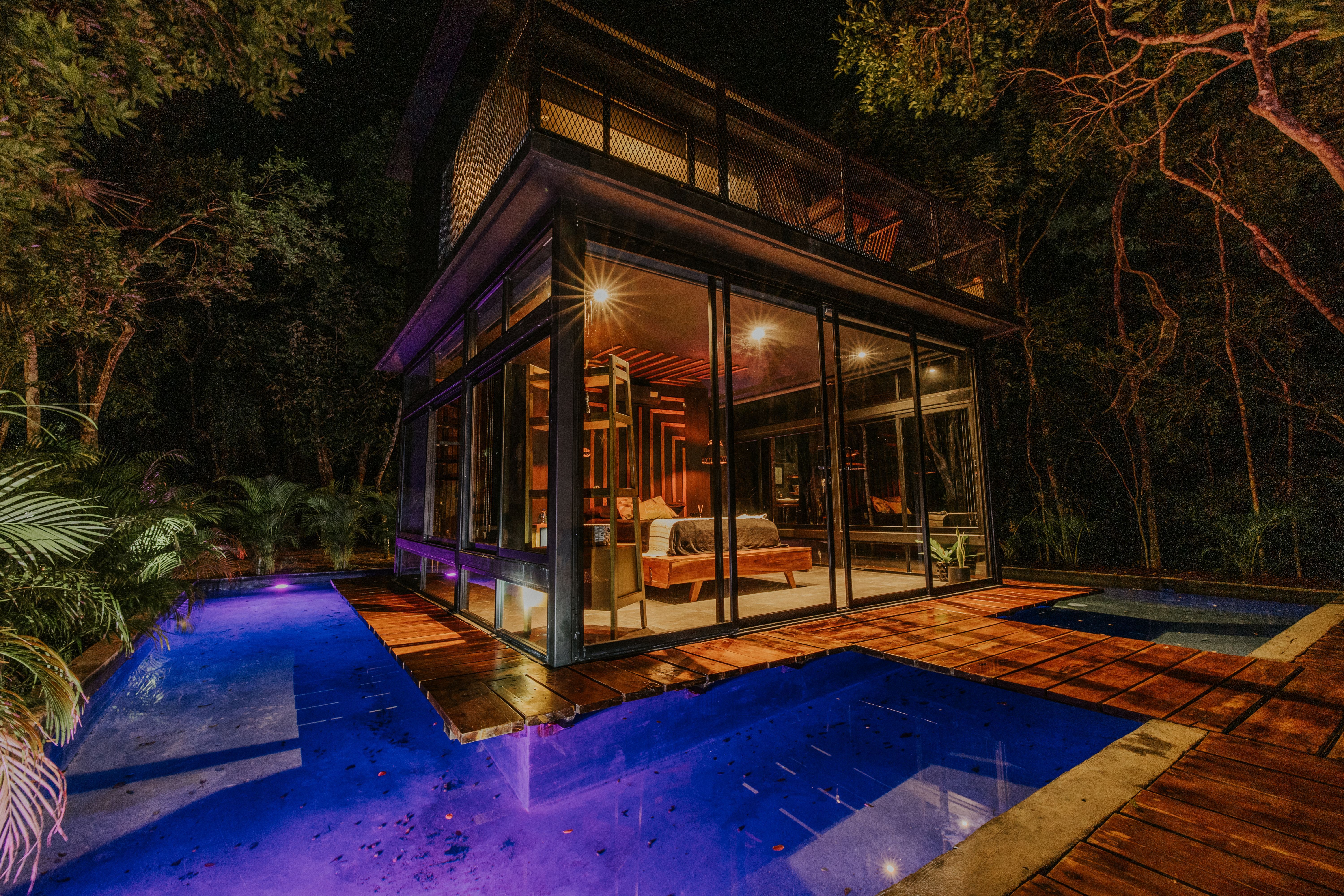 Suíte envidraçada iluminada à noite, com piscina refletindo luz entre a vegetação